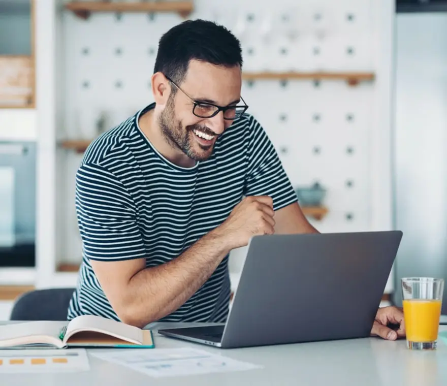 A smiling man looking at a laptop with an open book and a glass of orange juice.