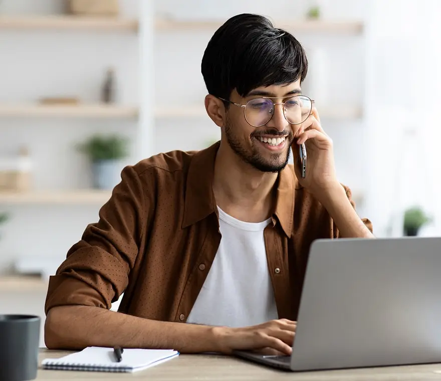 A smiling man is on the phone with customer service while looking at his laptop.